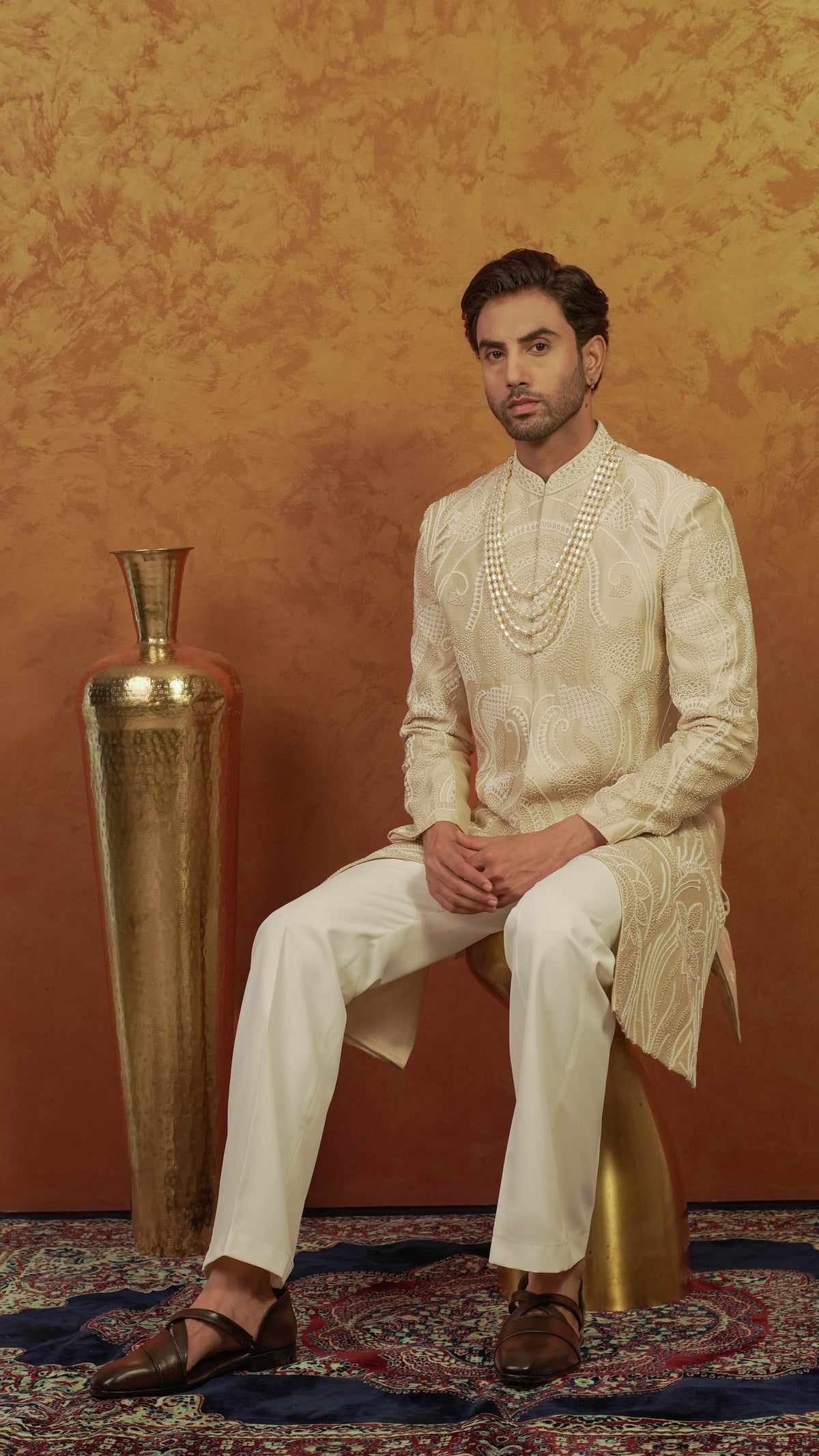 A man wearing a cream-colored embroidered sherwani and matching trousers sits on a gold stool against a textured golden-brown backdrop, with a large gold vase and patterned rug visible.
