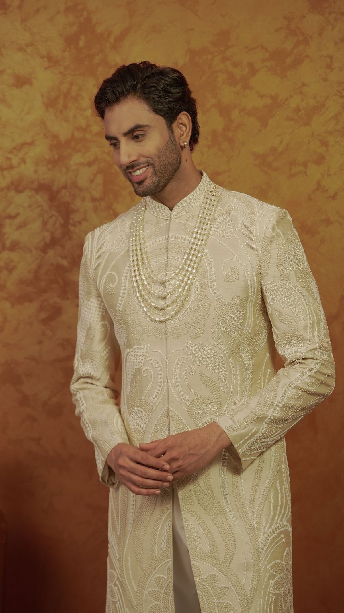 A man wearing a cream-colored embroidered sherwani with a matching double-row pearl necklace, standing against a textured golden-brown backdrop.