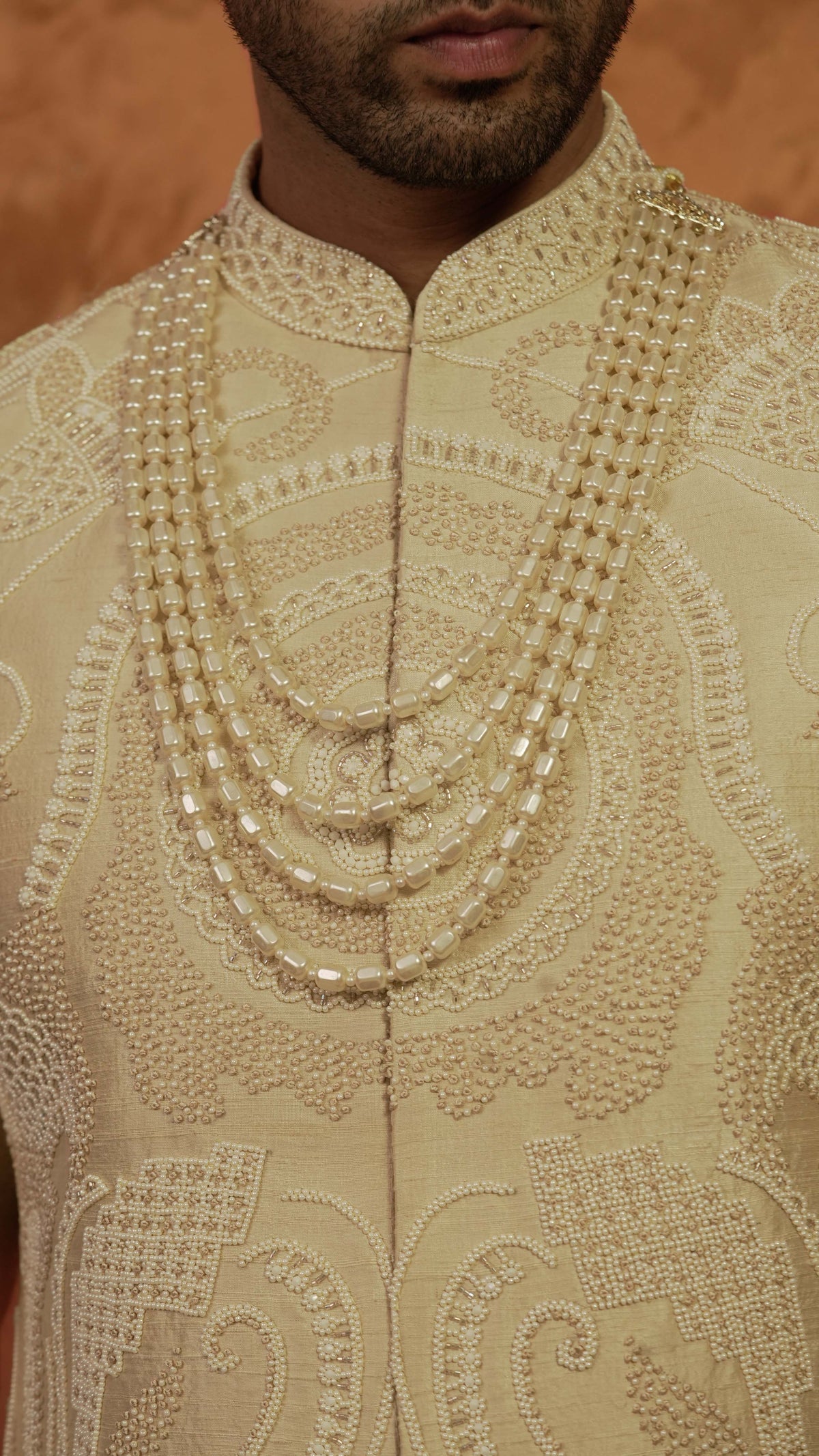 A close-up of a man wearing a cream-colored traditional sherwani with intricate embroidery and layered beaded necklaces.