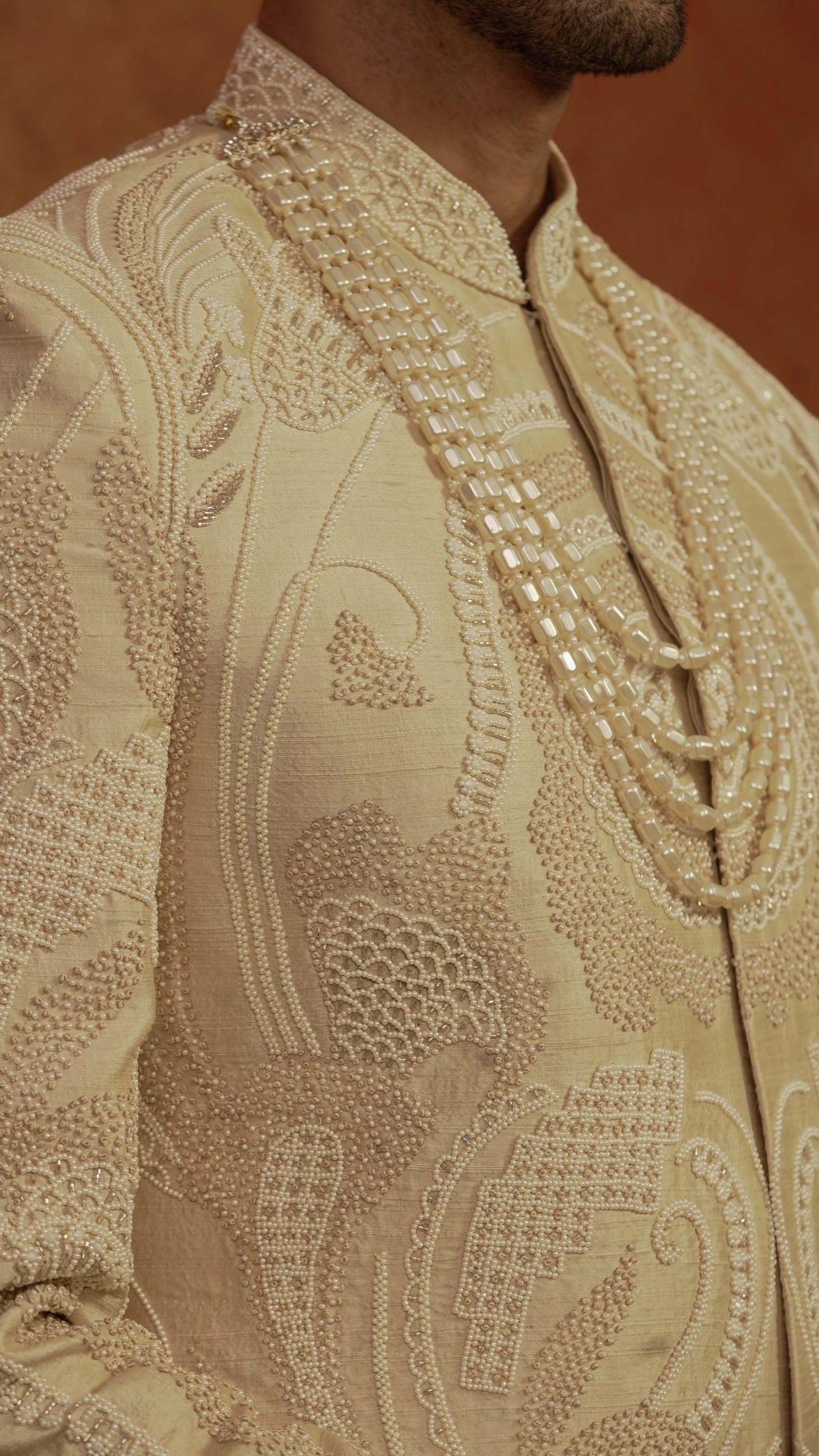 Close-up of a beige embroidered sherwani jacket with intricate metallic embroidery and a high collar, worn by a model.