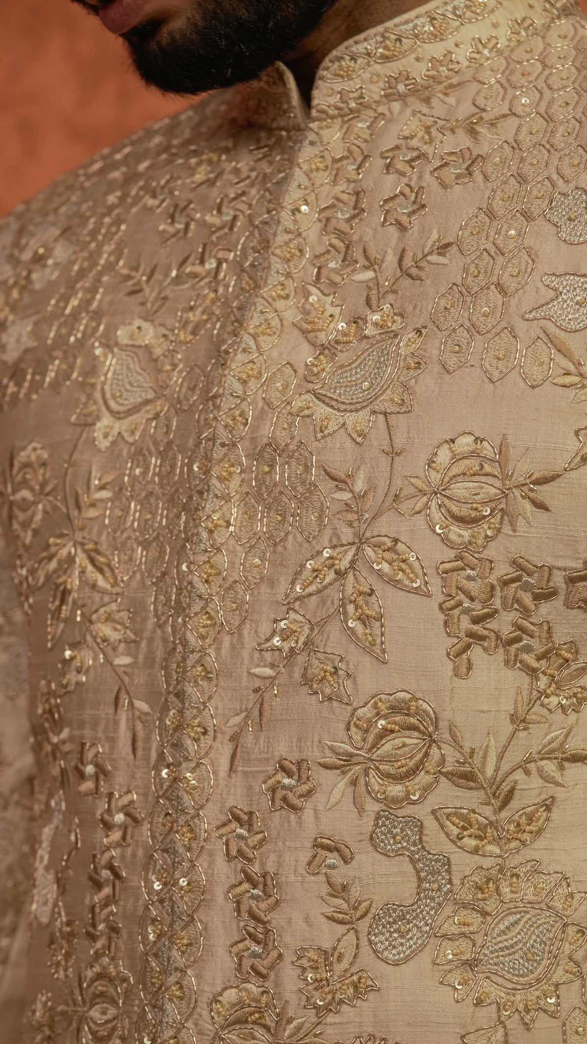 Close-up of a beige sherwani jacket with intricate gold floral embroidery and sequin detailing, worn by a man.