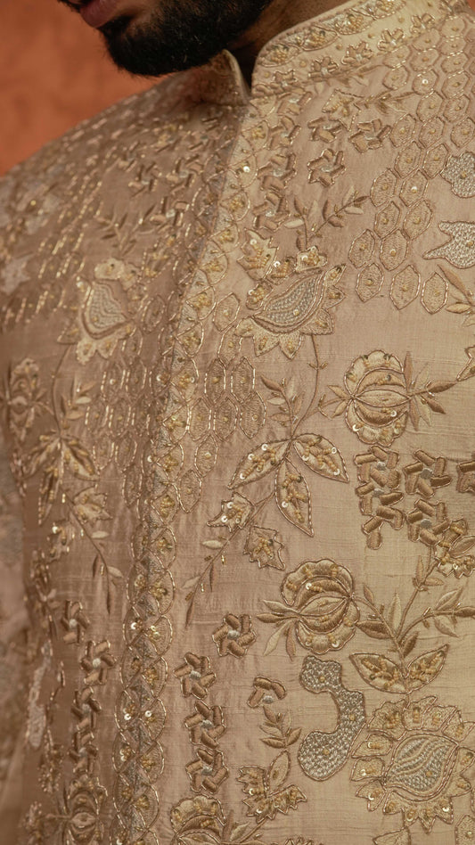Close-up of a beige sherwani jacket with intricate gold floral embroidery and sequin detailing, worn by a man.