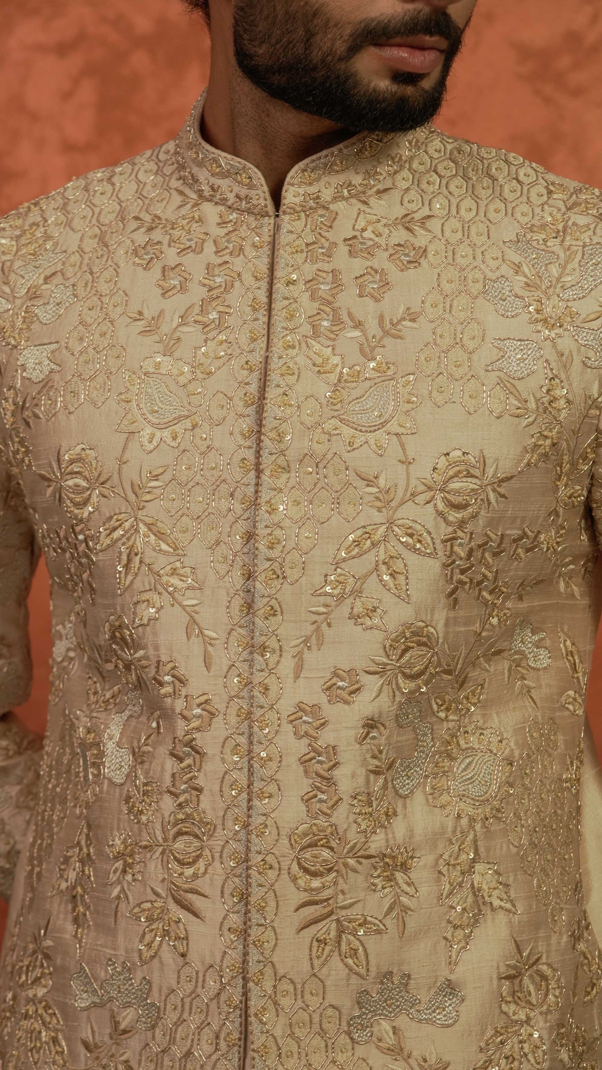 A close-up of a man wearing a beige sherwani jacket with intricate gold and silver floral embroidery, set against a warm orange background.