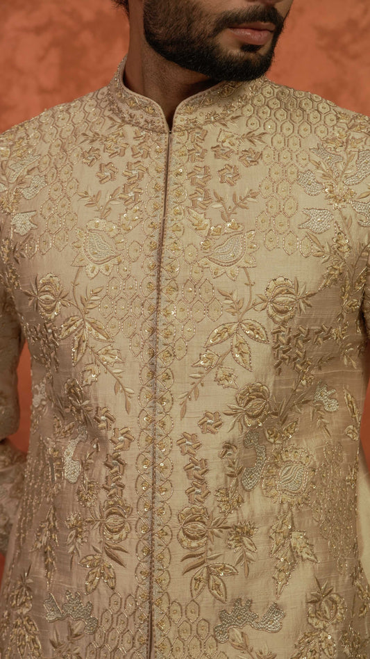 A close-up of a man wearing a beige sherwani jacket with intricate gold and silver floral embroidery, set against a warm orange background.
