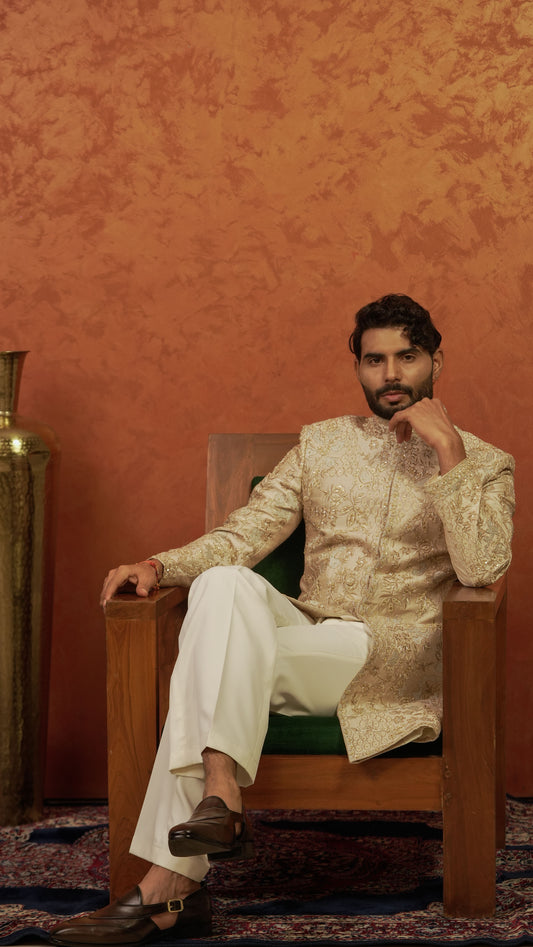 A man wearing a traditional Indian outfit with a beige and gold embroidered sherwani and white trousers, sitting in a wooden armchair against a textured orange wall.