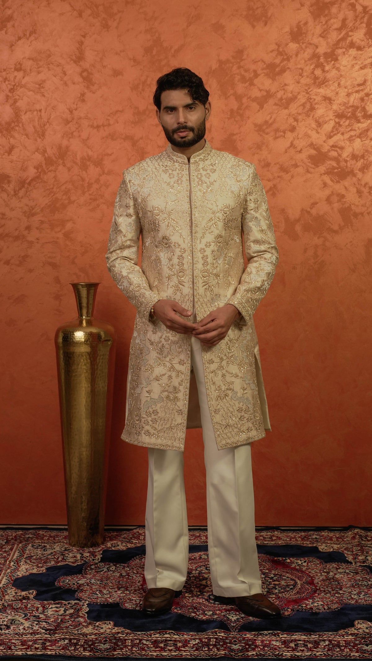 A man wearing a cream and gold embroidered sherwani jacket with matching cream trousers, standing in front of a textured orange wall next to a gold decorative vase.