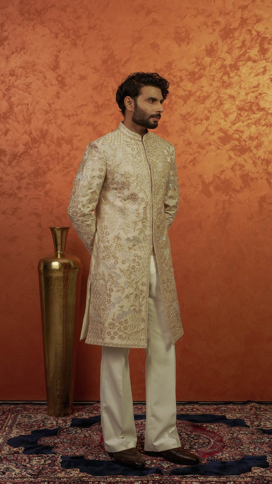 A man wearing a traditional Indian outfit, featuring a cream-colored embroidered sherwani jacket and matching trousers, standing in front of a textured orange wall next to a gold vase.