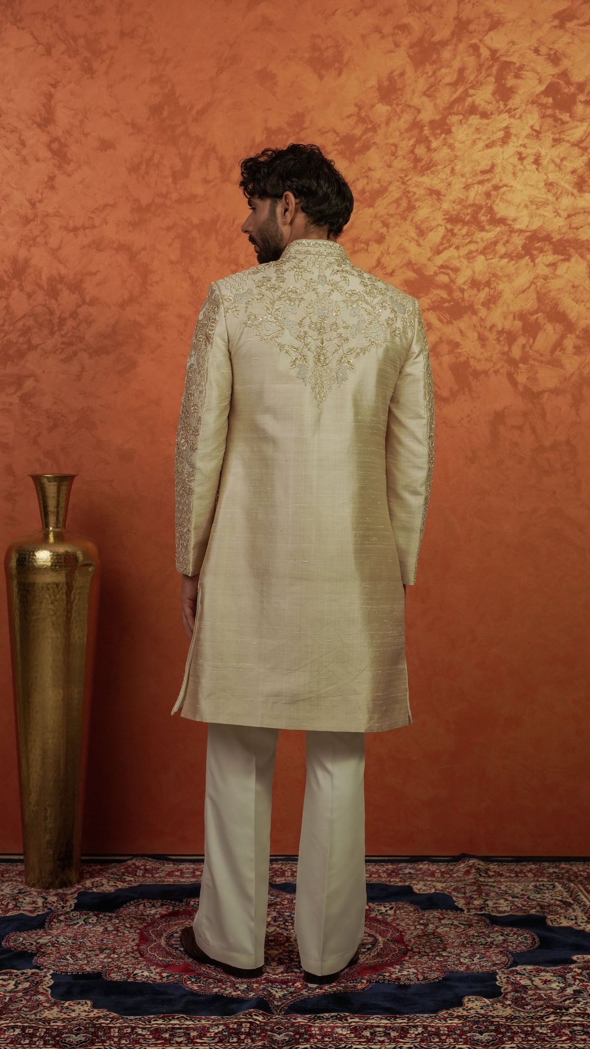 A man viewed from behind wearing a cream-colored embroidered sherwani jacket with matching trousers, standing on a patterned rug against a textured orange wall.