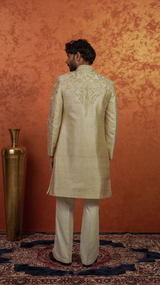 A man viewed from behind wearing a cream-colored embroidered sherwani jacket with matching trousers, standing on a patterned rug against a textured orange wall.