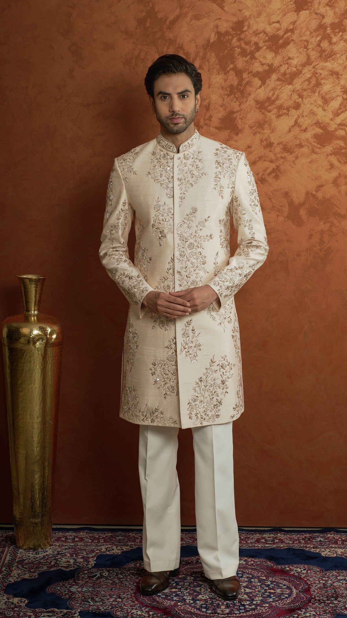 A man wearing a light rose gold-colored sherwani jacket with intricate gold embroidery and matching cream trousers, standing against a textured orange-brown backdrop next to a gold decorative vase.