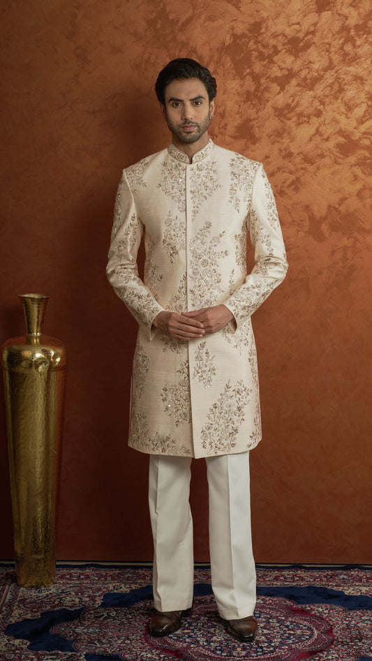 A man wearing a light rose gold-colored sherwani jacket with intricate gold embroidery and matching cream trousers, standing against a textured orange-brown backdrop next to a gold decorative vase.