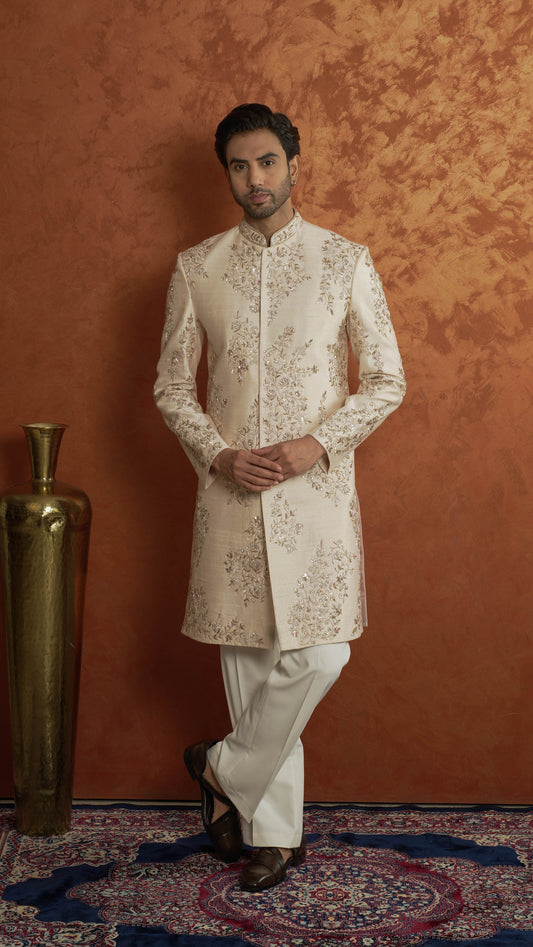 A man wearing a light rose gold sherwani with intricate gold embroidery and white trousers, standing against a textured orange-brown wall next to a gold decorative vase.