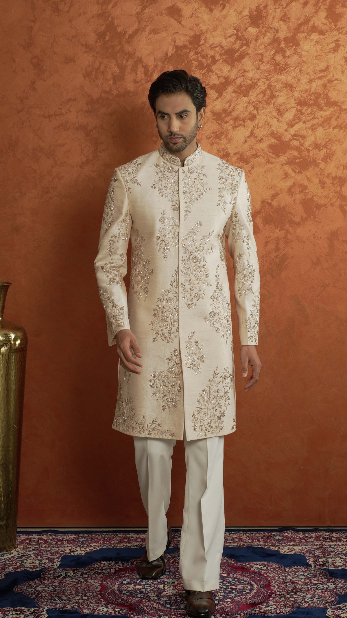 A man modeling a traditional light rose gold sherwani with intricate gold embroidery, paired with matching cream trousers, set against a textured orange-brown studio backdrop.