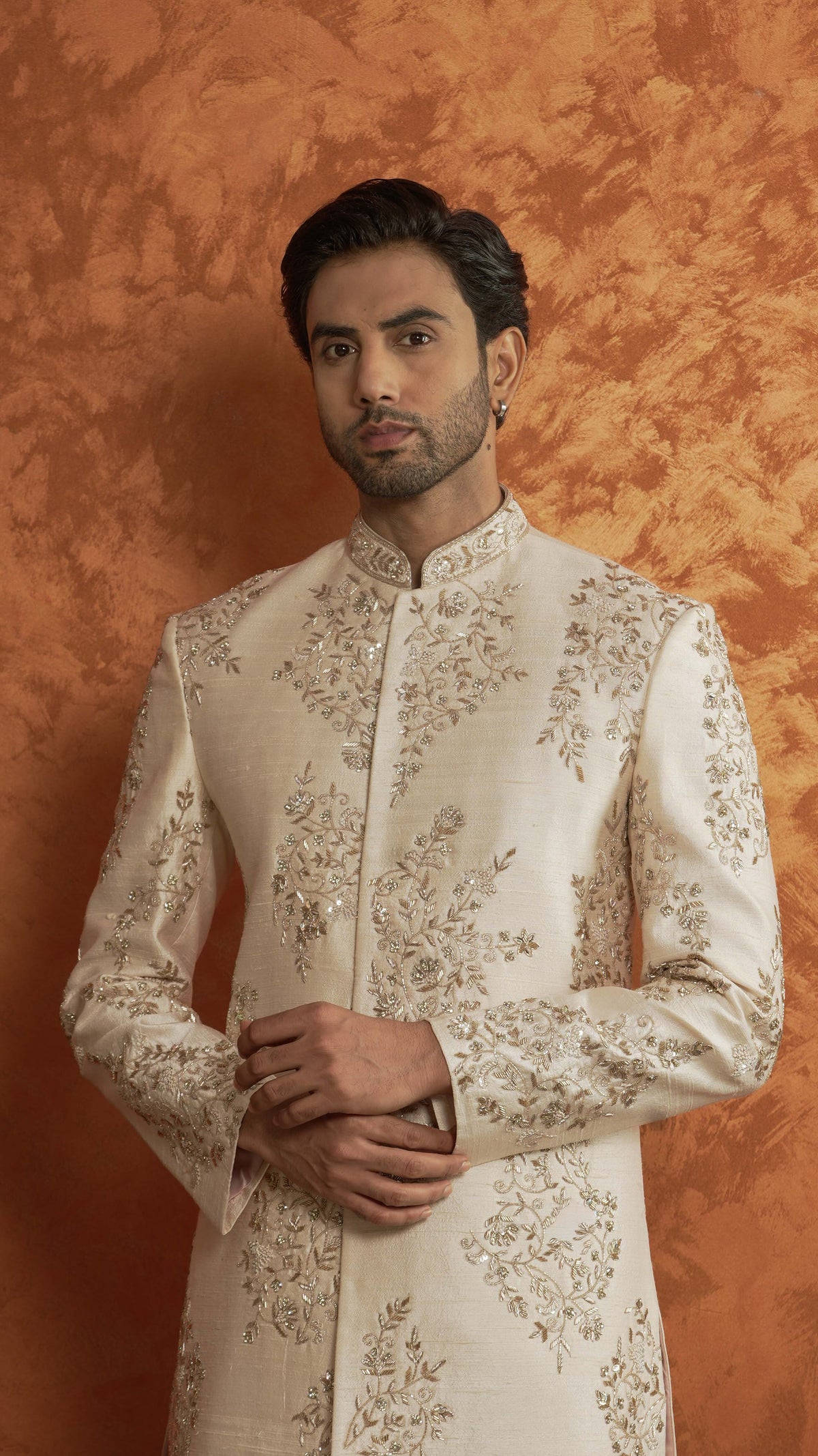 A man wearing a light rose gold-colored traditional sherwani jacket with intricate gold floral embroidery, standing against a textured orange-brown backdrop.