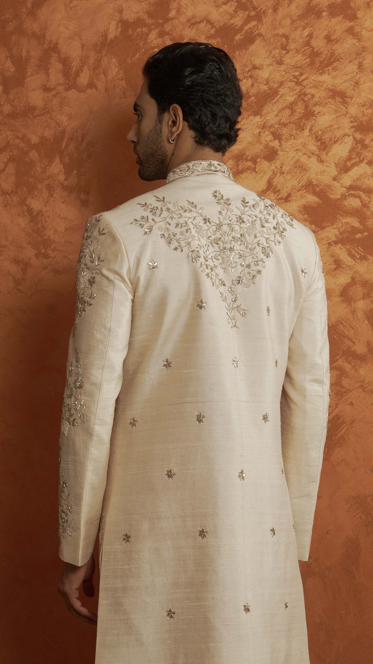 A man shown from the back wearing a light rose gold-colored traditional sherwani with intricate gold embroidery on the shoulders and scattered star motifs, set against a textured orange-brown backdrop.