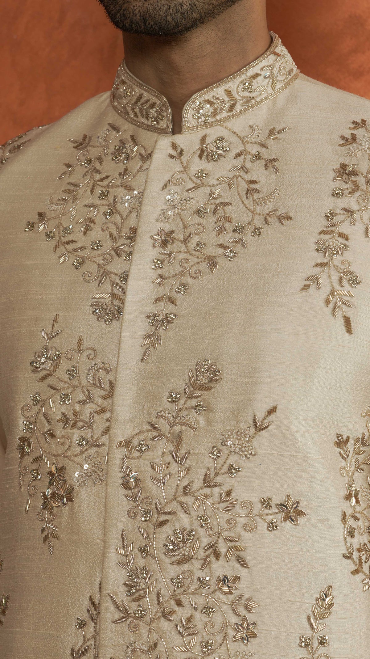 Close-up of a light rose gold sherwani jacket with intricate silver and gold floral embroidery and beadwork, worn by a model against a warm brown background.