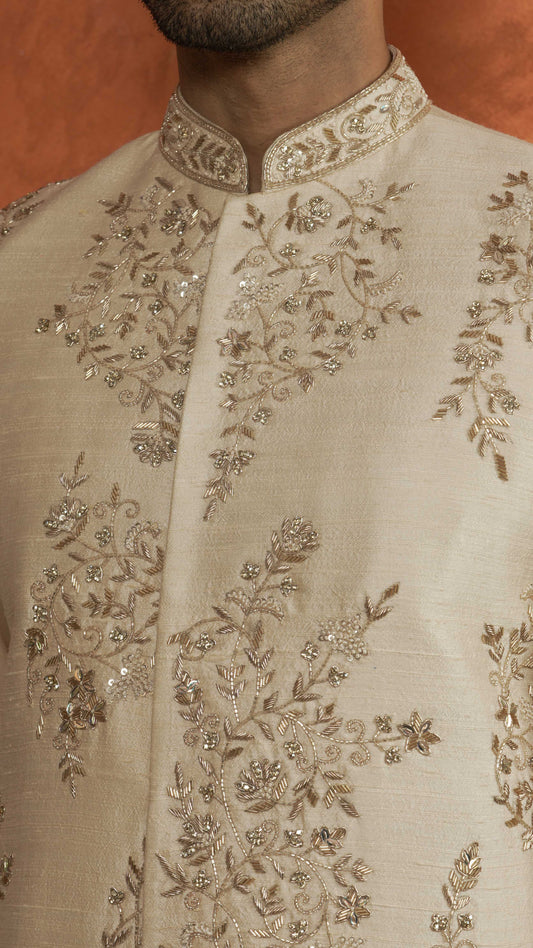 Close-up of a light rose gold sherwani jacket with intricate silver and gold floral embroidery and beadwork, worn by a model against a warm brown background.