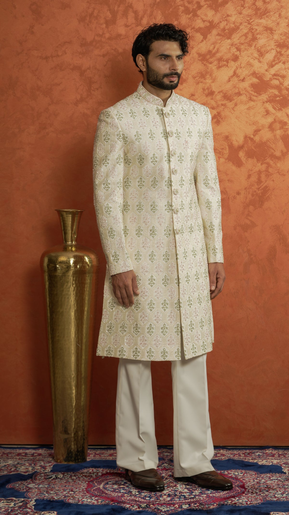 A man modeling a traditional Indian outfit consisting of a cream-colored sherwani jacket with light green floral patterns, paired with cream trousers, standing against a textured orange wall next to a gold decorative vase.