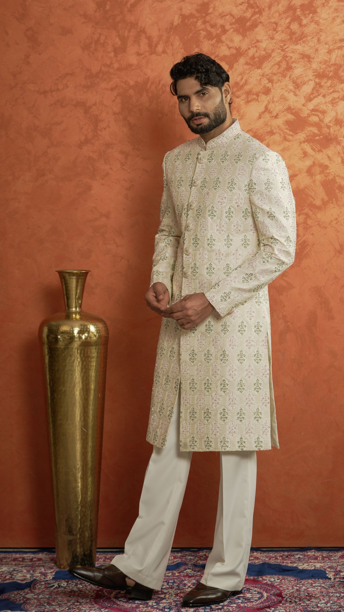 A man modeling a traditional Indian outfit consisting of a cream-colored sherwani with light green motifs and white trousers, standing in front of a textured orange wall next to a large gold decorative vase.
