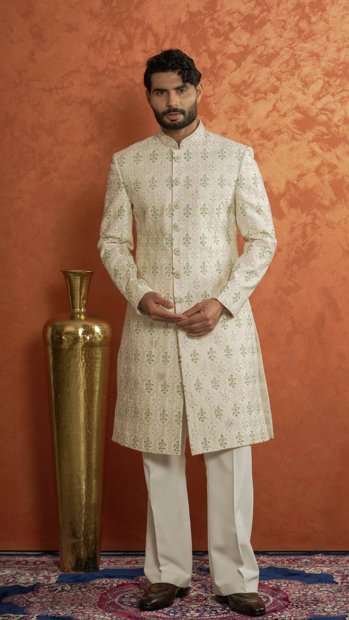 A man wearing a cream sherwani with green floral motifs and white trousers, standing in front of a textured orange wall next to a large gold decorative vase.