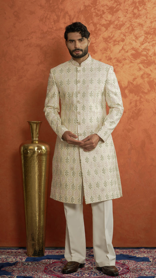 A man wearing a cream sherwani with green floral motifs and white trousers, standing in front of a textured orange wall next to a large gold decorative vase.