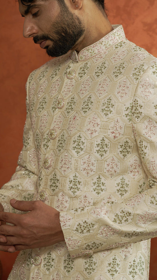 A close-up of a man wearing a cream-colored traditional Indian sherwani jacket with intricate green and pink floral embroidery and gold buttons, set against a warm orange background.