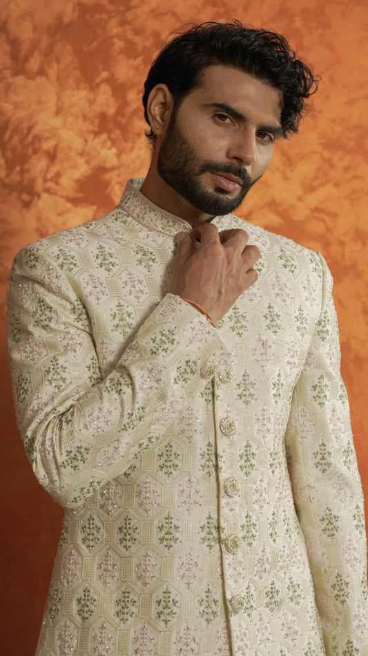 A man wearing a cream-colored sherwani jacket with green and pink floral patterns, posed against a textured orange background.