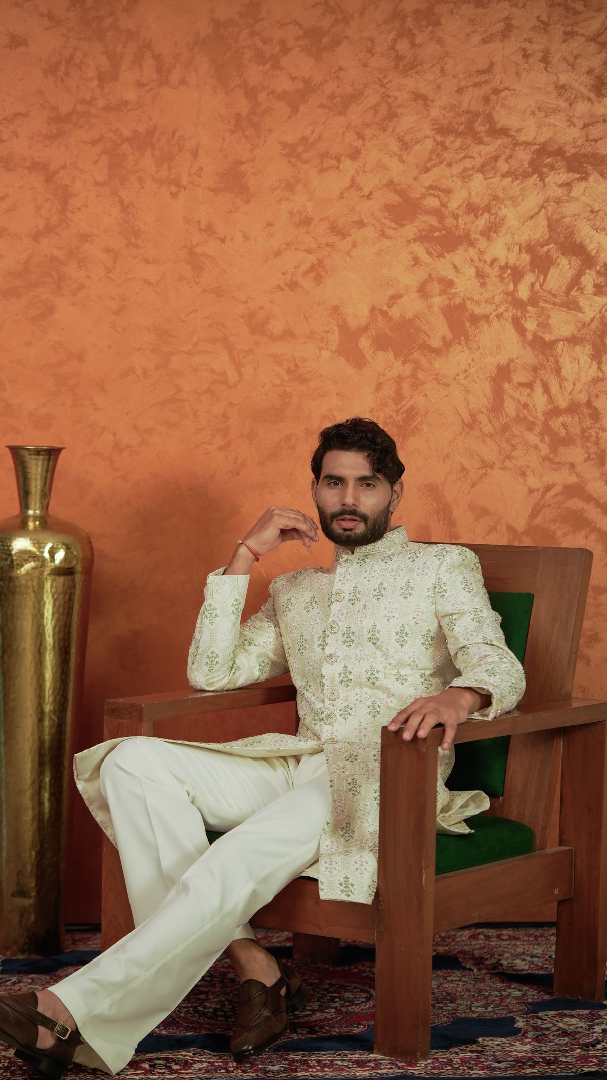 A man wearing a cream-colored embroidered sherwani and matching trousers sits in a wooden armchair with a green cushion against a textured orange wall.