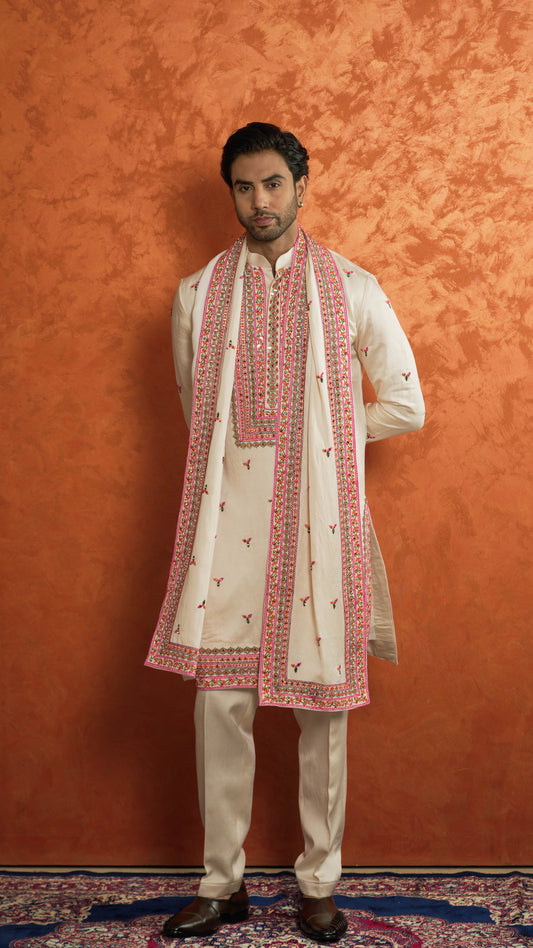 A man modeling a traditional Indian outfit consisting of a cream-colored kurta with pink and gold embroidery, matching trousers, and a long cream shawl with pink borders and embroidery, set against a textured orange backdrop.