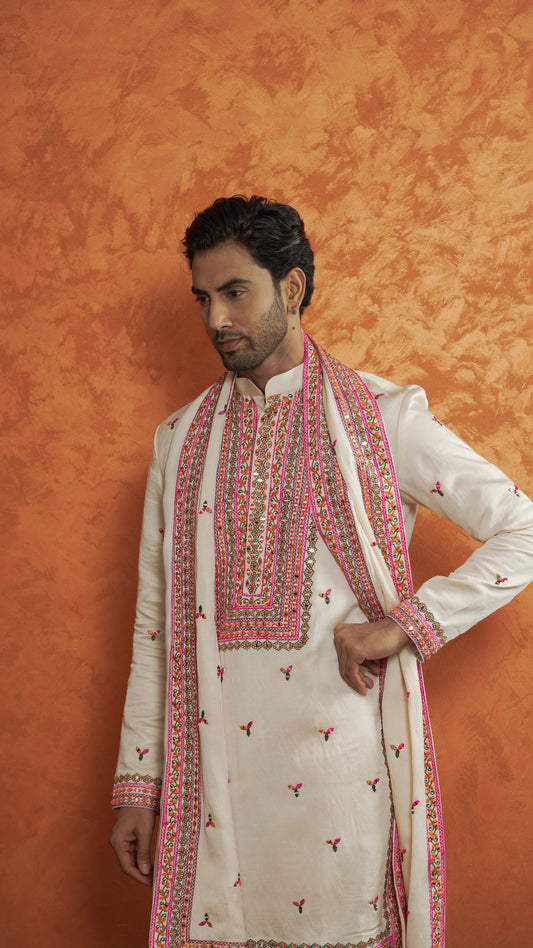 A man wearing a traditional white Indian kurta with pink and gold embroidery, paired with a matching embroidered dupatta, posed against a textured orange background.