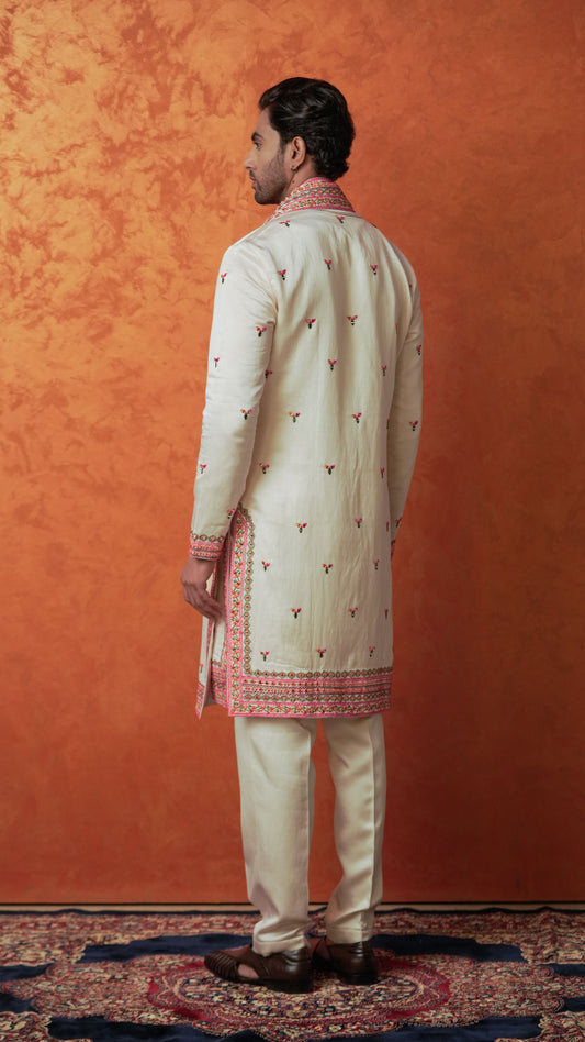 A man wearing a cream-colored traditional multicolor kurta and trousers set with orange green embroidery, shown from the back against a textured orange backdrop.
