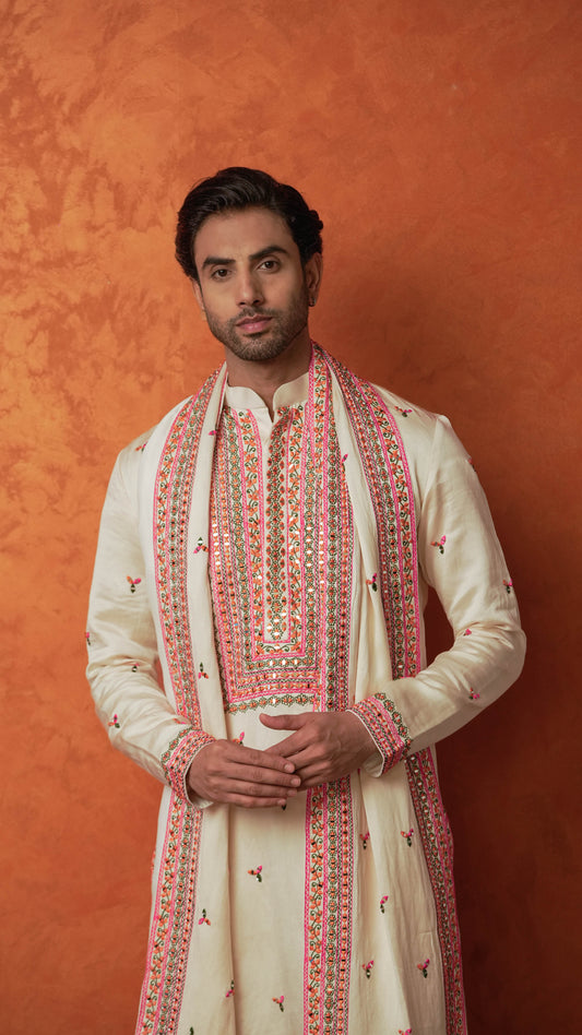 A man wearing a cream-colored traditional Indian outfit with pink and orange embroidery, standing against a textured orange background.