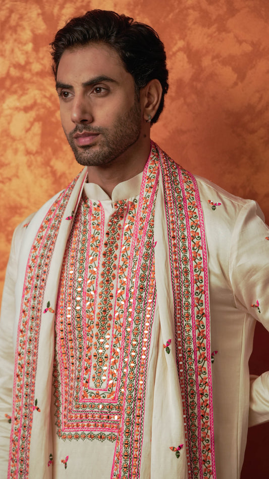 A man wearing a cream-colored traditional Indian outfit with a matching embroidered sherwani and a pink -orange embroidered scarf, set against a textured orange background.