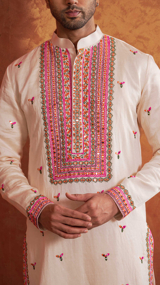 A man wearing a cream-colored kurta with vibrant pink, orange, and green embroidery around the collar and cuffs, posed against a textured orange-brown background.