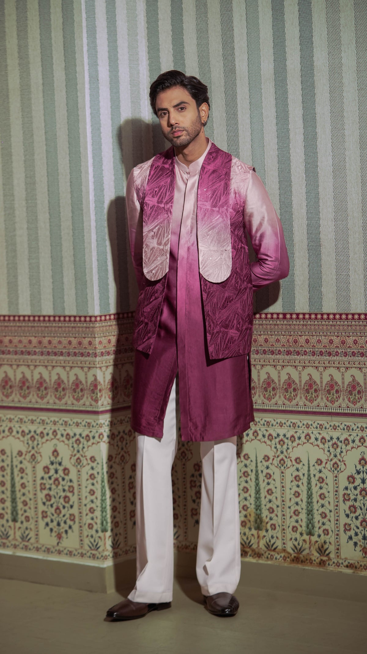 A man modeling a traditional Indian outfit consisting of a magenta and pink gradient embroidered vest over a white shirt, paired with white trousers and dark brown shoes, standing against a patterned wallpaper background.