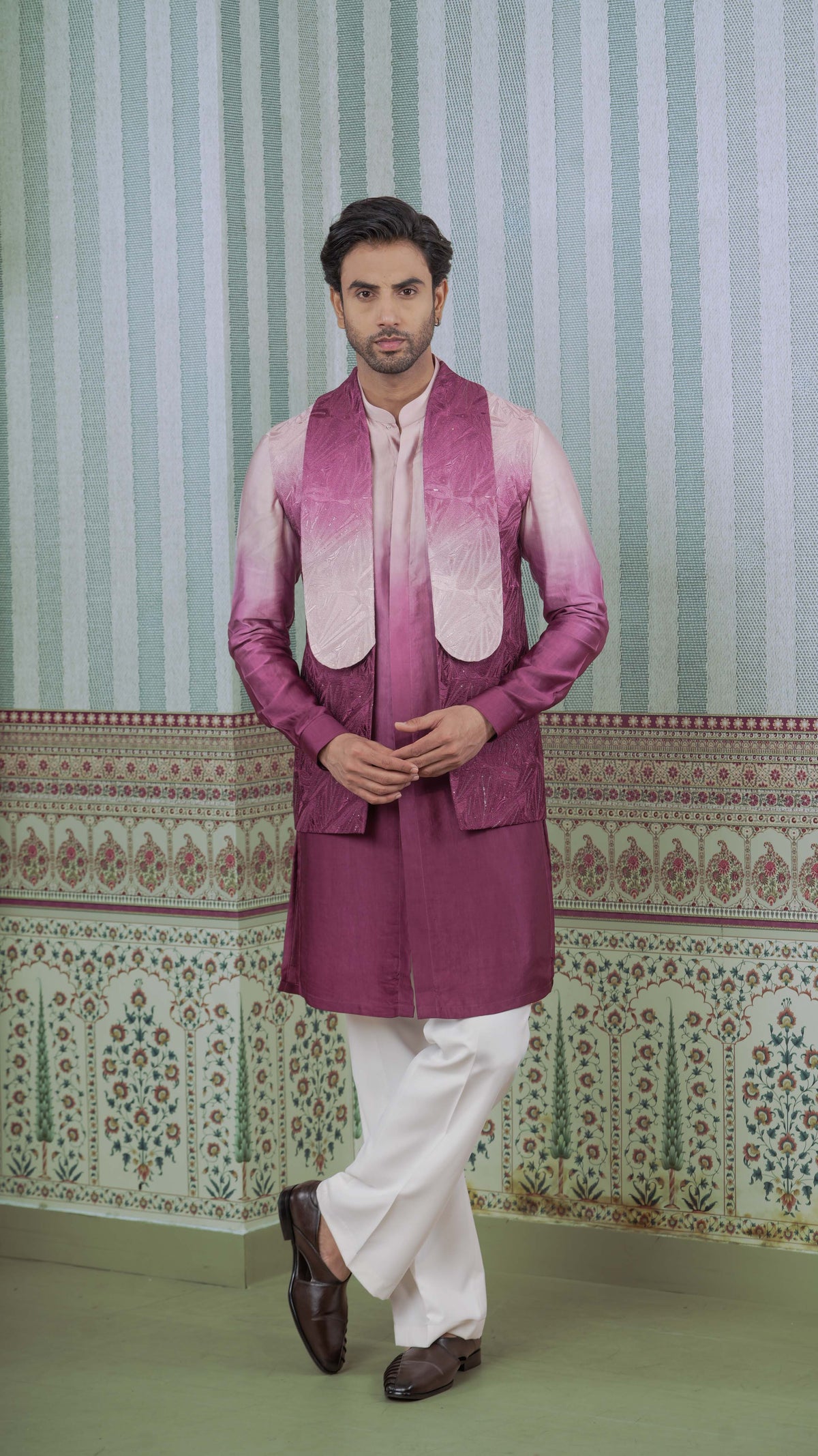 A man modeling a traditional Indian outfit featuring a magenta ombre jacket over a ombre pink kurta, paired with white trousers and dark brown loafers, set against a patterned green and white wall.