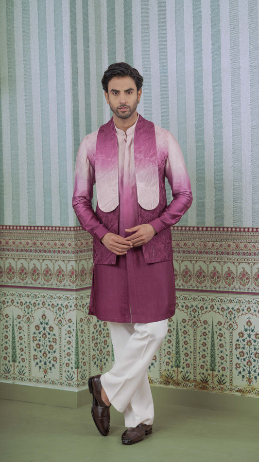 A man modeling a traditional Indian outfit featuring a magenta ombre jacket over a ombre pink kurta, paired with white trousers and dark brown loafers, set against a patterned green and white wall.