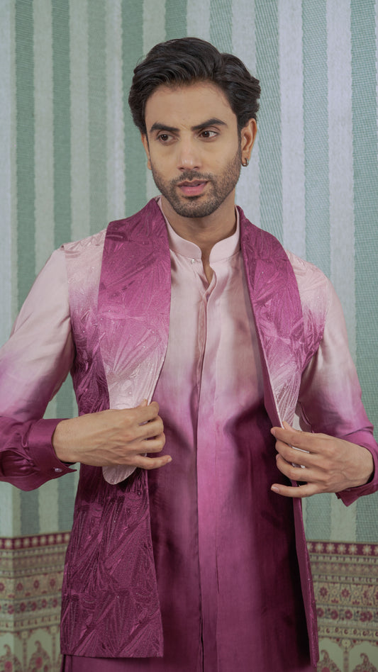 A man wearing a traditional Indian outfit consisting of a light pink kurta and a magenta-purple satin vest, standing in front of a green and white striped background.