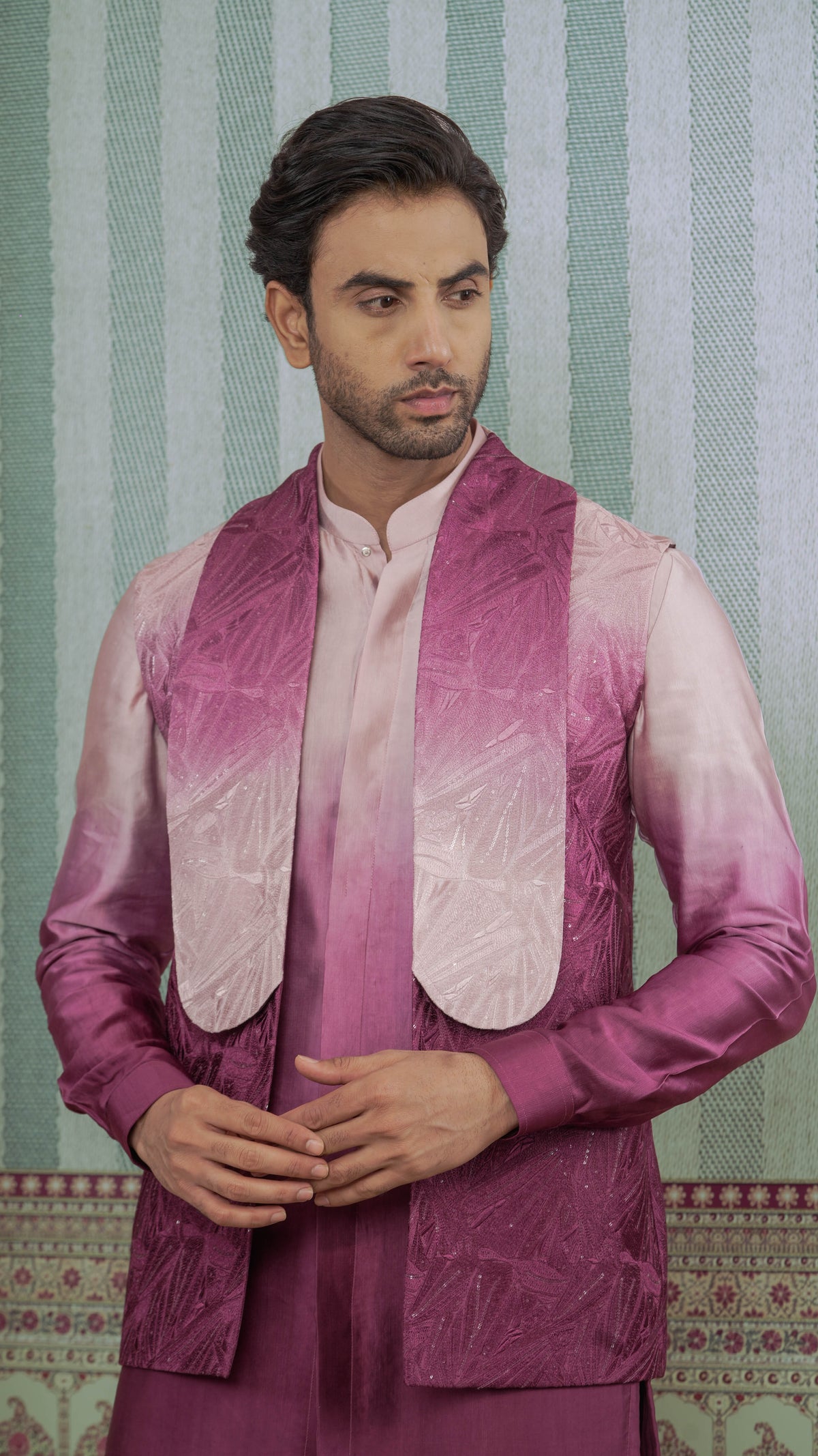 A man wearing a traditional Indian outfit featuring a magenta and pink gradient sherwani jacket over a light pink kurta, set against a green and white striped patterned backdrop.