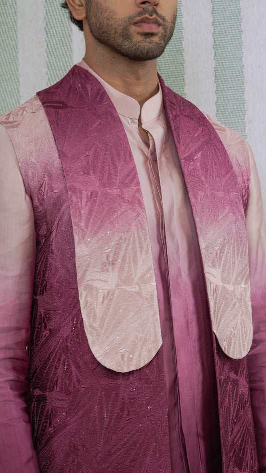 A close-up of a man wearing a traditional Indian outfit featuring a magenta and pink gradient embroidered vest over a light pink kurta, with a textured green and white striped background.