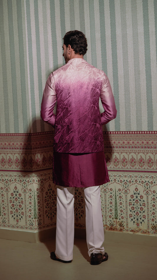 A man viewed from behind wearing a metallic magenta and purple gradient embroidered kurta with white trousers, standing in front of a green and white striped wallpapered wall.