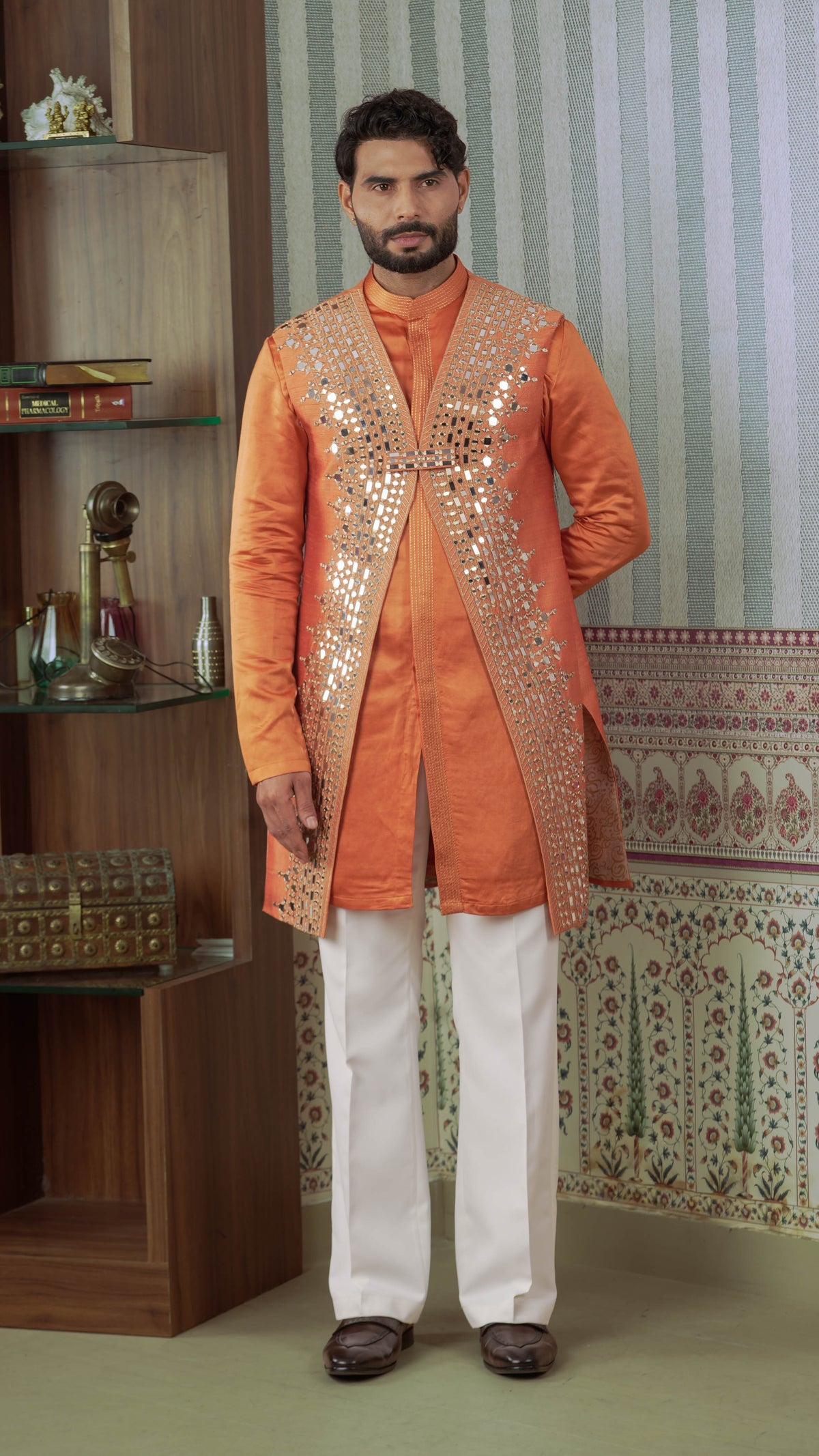 A man wearing a traditional Indian outfit consisting of an orange kurta with silver mirror work embellishments, white trousers, and brown shoes, standing in an indoor setting with a patterned wall and wooden shelves.