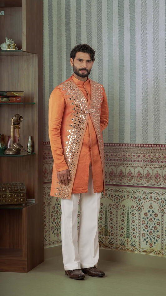 A man modeling a traditional Indian outfit consisting of an orange kurta with silver mirror work, white trousers, and brown leather shoes, standing in front of a patterned wall.