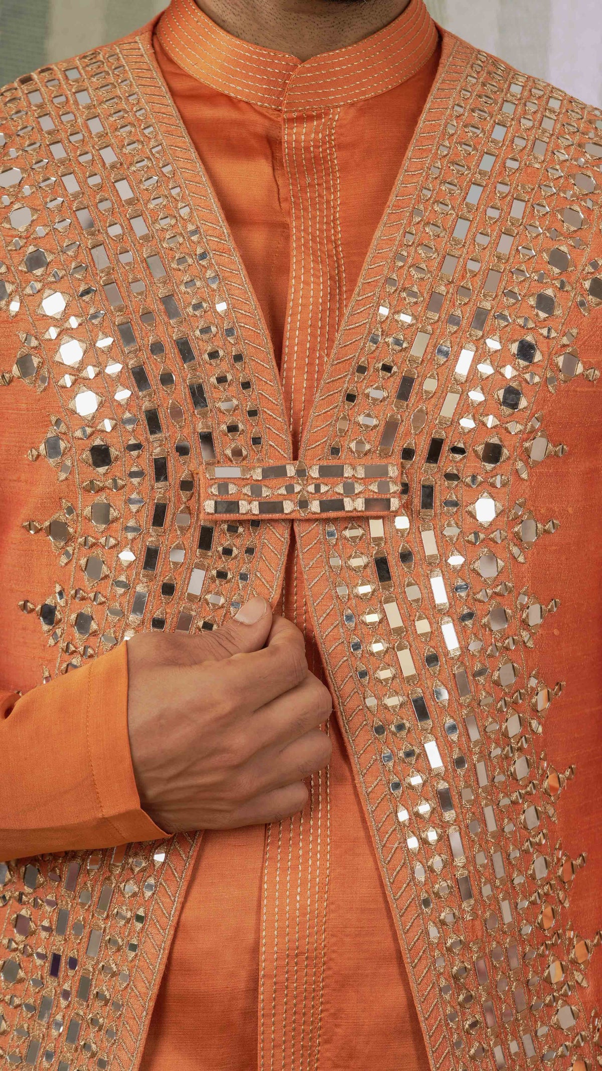 Close-up of a person wearing an orange kurta with extensive silver mirror work and metallic embellishments on the front panel and lapels.