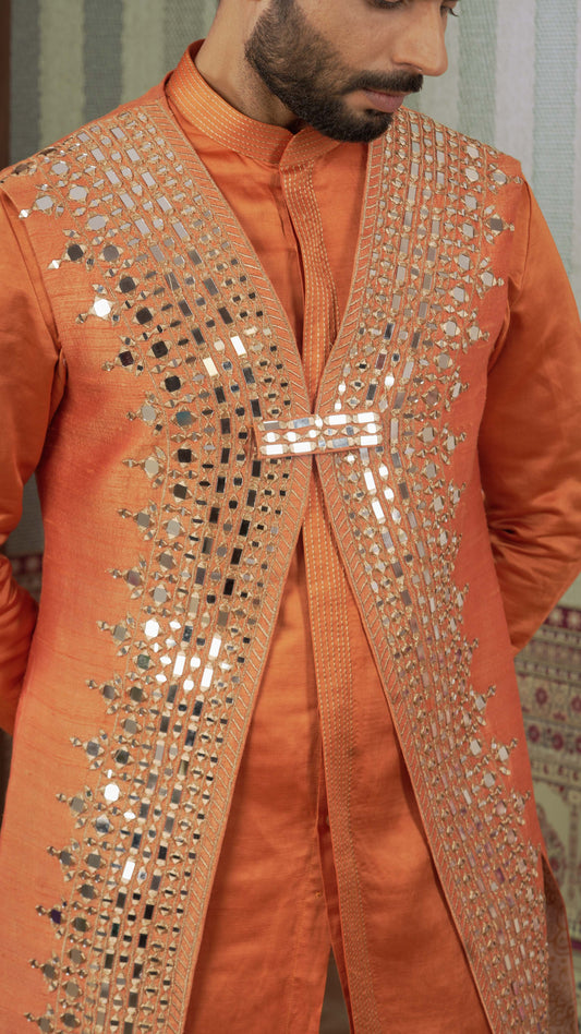 A close-up of a man wearing a bright orange traditional outfit with a matching embroidered vest featuring extensive silver mirror work and geometric patterns.