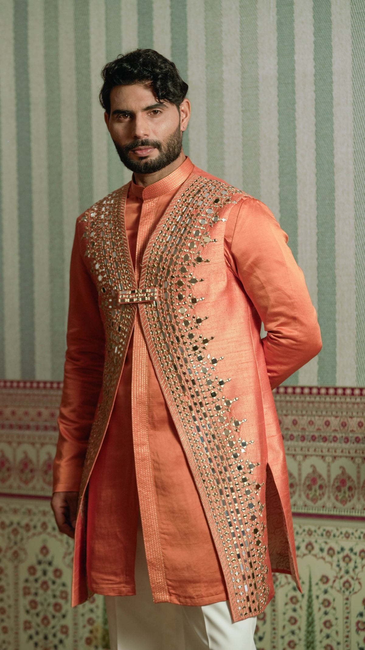 A man wearing a peach-colored traditional Indian kurta with extensive gold and silver mirror work embroidery on the front, paired with white trousers, standing against a green and white patterned wallpaper background.
