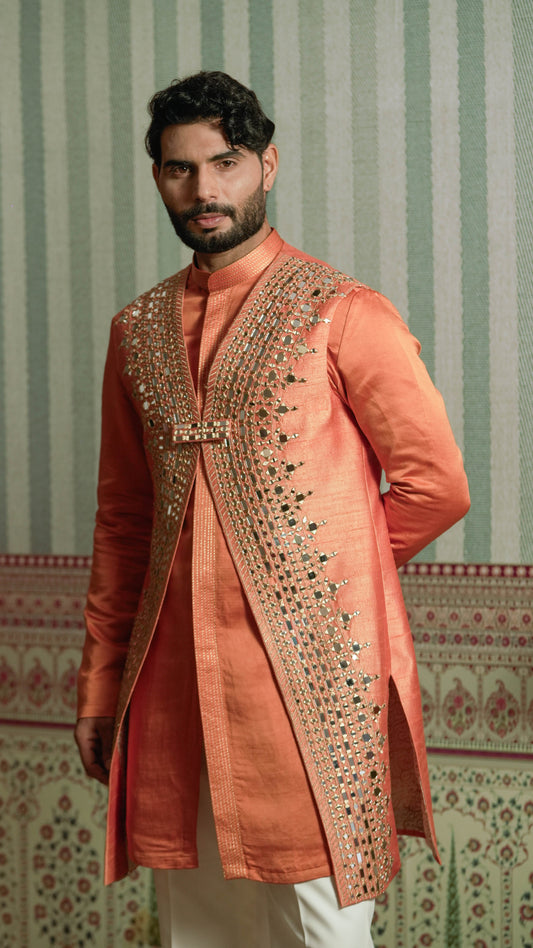 A man wearing a peach-colored traditional Indian kurta with extensive gold and silver mirror work embroidery on the front, paired with white trousers, standing against a green and white patterned wallpaper background.