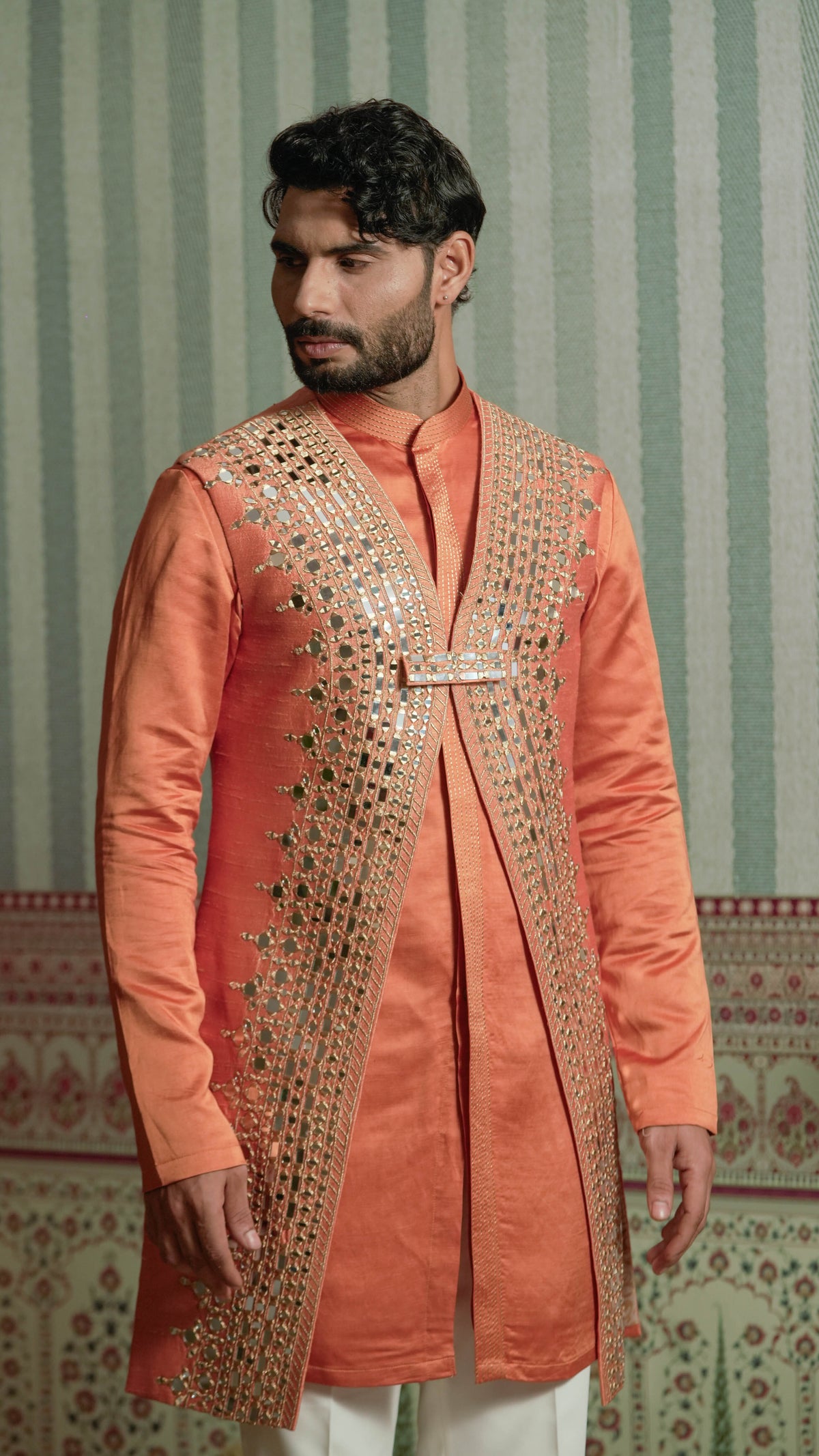 A man wearing a peach-colored traditional kurta with ornate gold mirror work embroidery on the front, paired with white trousers, standing against a green and white patterned wallpaper background.