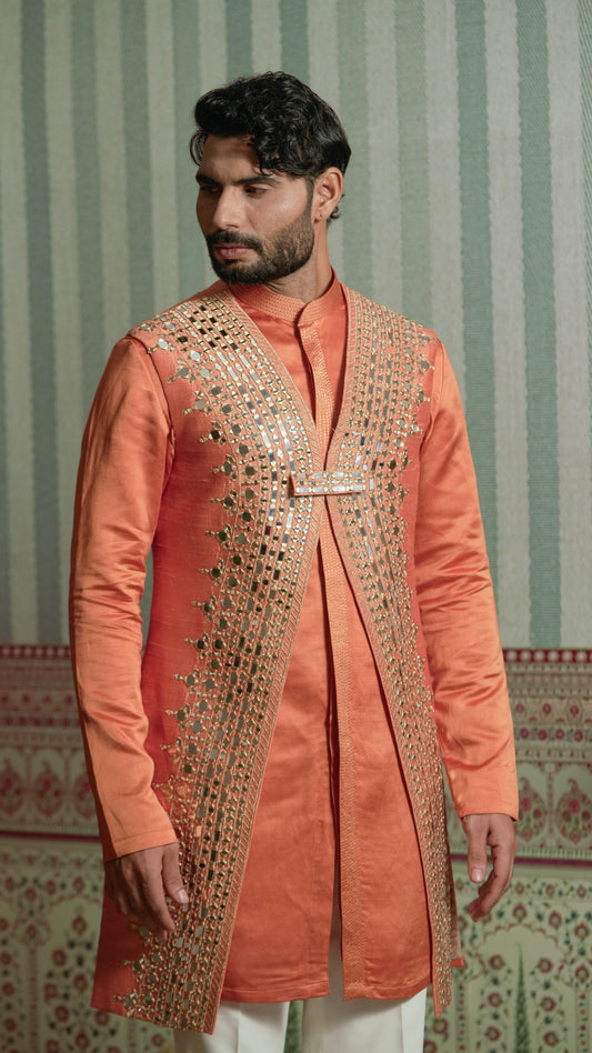 A man wearing a peach-colored traditional kurta with ornate gold mirror work embroidery on the front, paired with white trousers, standing against a green and white patterned wallpaper background.