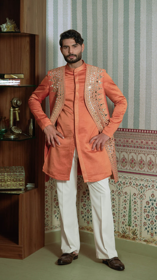 A man modeling a peach-orange kurta with gold embroidery on the front, paired with white trousers and brown shoes, standing in front of a patterned wallpaper and wooden shelf.
